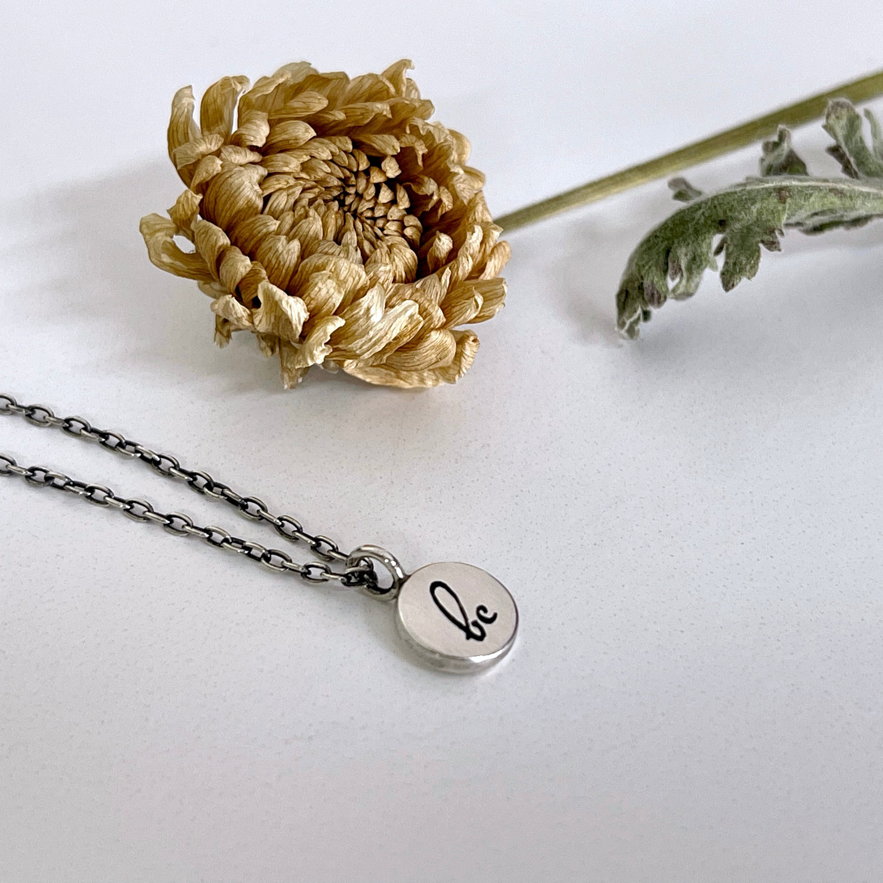 "be" charm necklace shown at an angle next to a dried flower and leaf