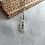 Silver necklace with a rectangular pendant engraved with 'PAPA' on a light fabric background.