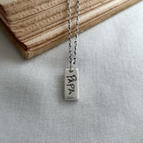 Silver necklace with a rectangular pendant engraved with 'PAPA' on a light fabric background.