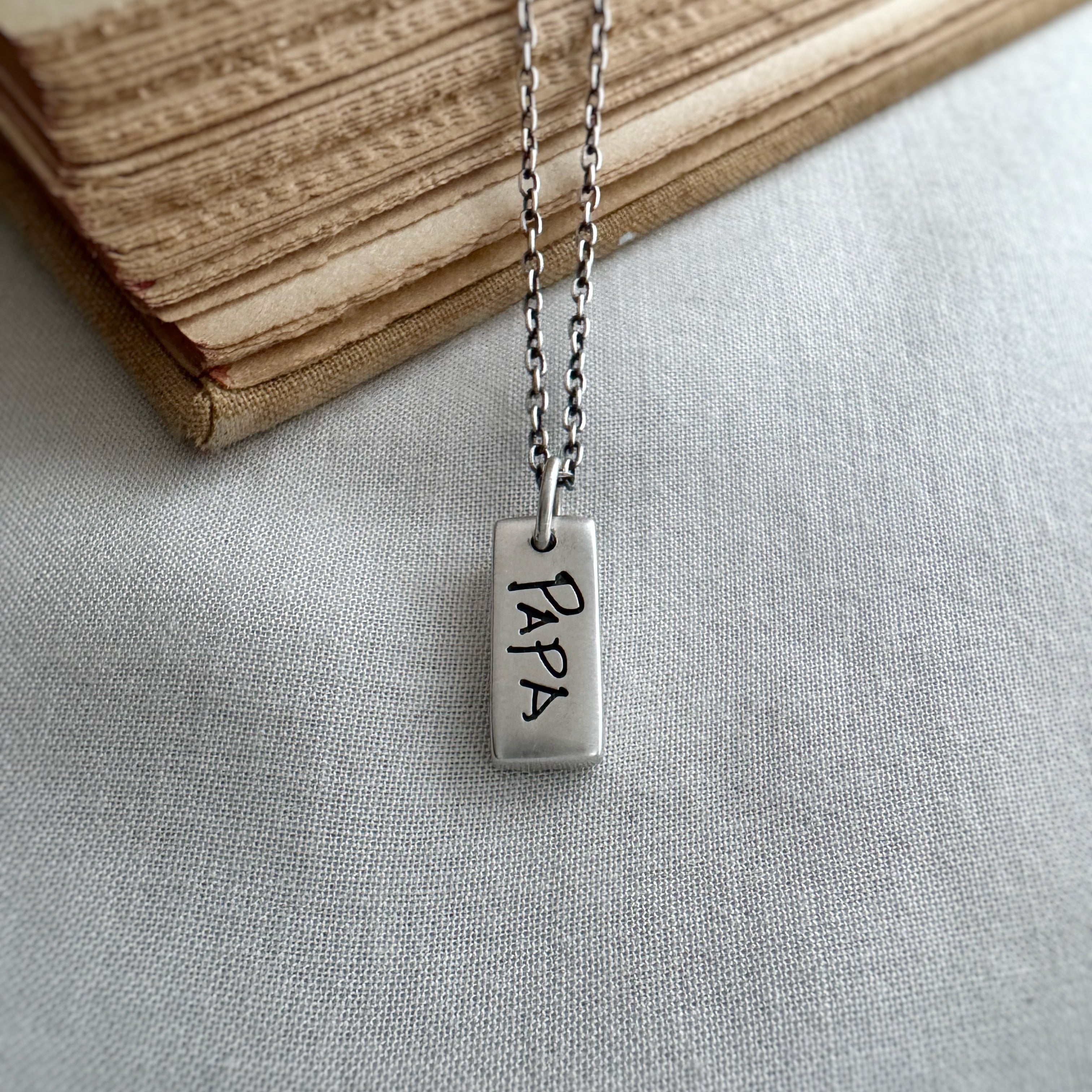 Silver necklace with a rectangular pendant engraved with 'PAPA' on a light fabric background.