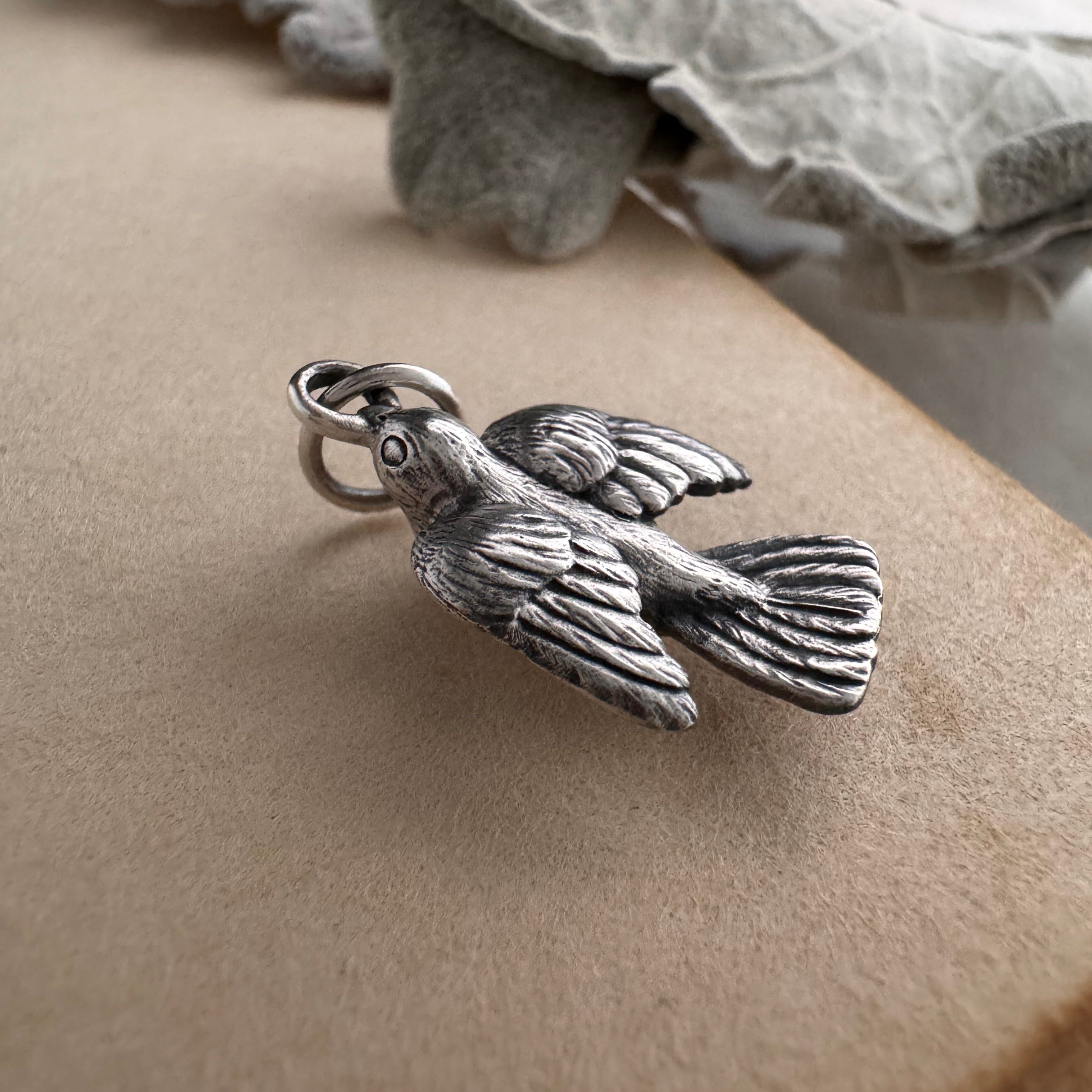 Silver sparrow charm sits on an old book with leaves in the background.  Each feather in the bird's wings and tail are intricately carved with great detail.