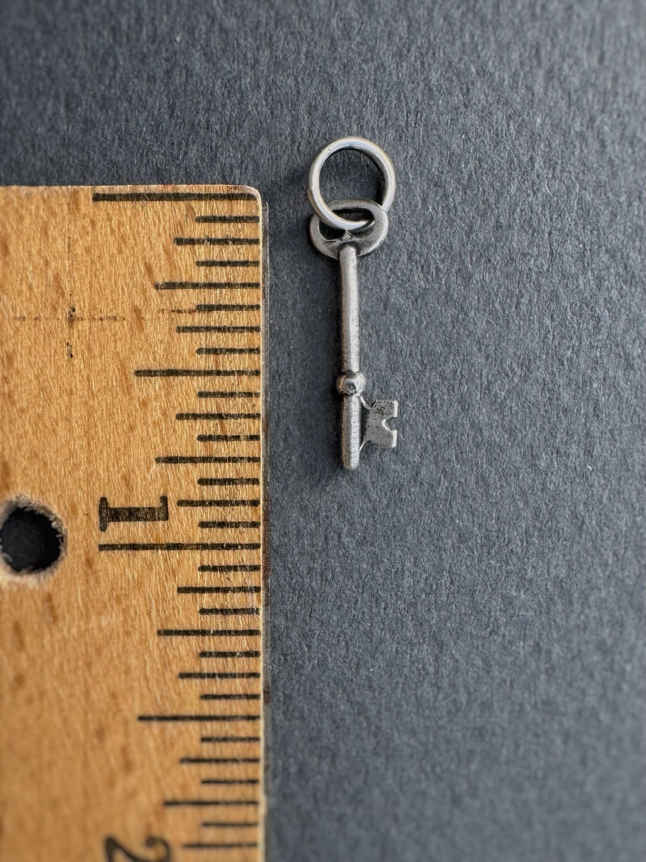 Small silver key charm next to a wooden ruler on a gray background. Key measures 3/4" long