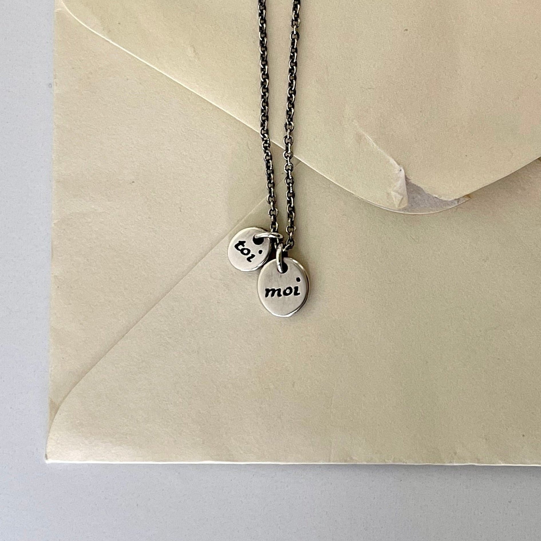 A silver necklace with two small inscribed charms attached to a beige envelope.