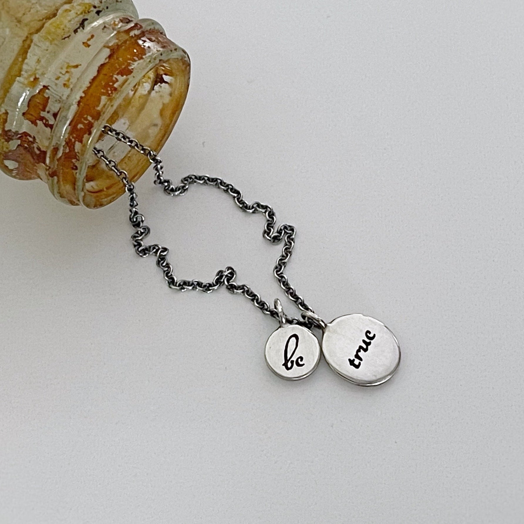 A silver necklace with two round charms that say 'BE TRUE' on a white background, attached to a glass bottle.