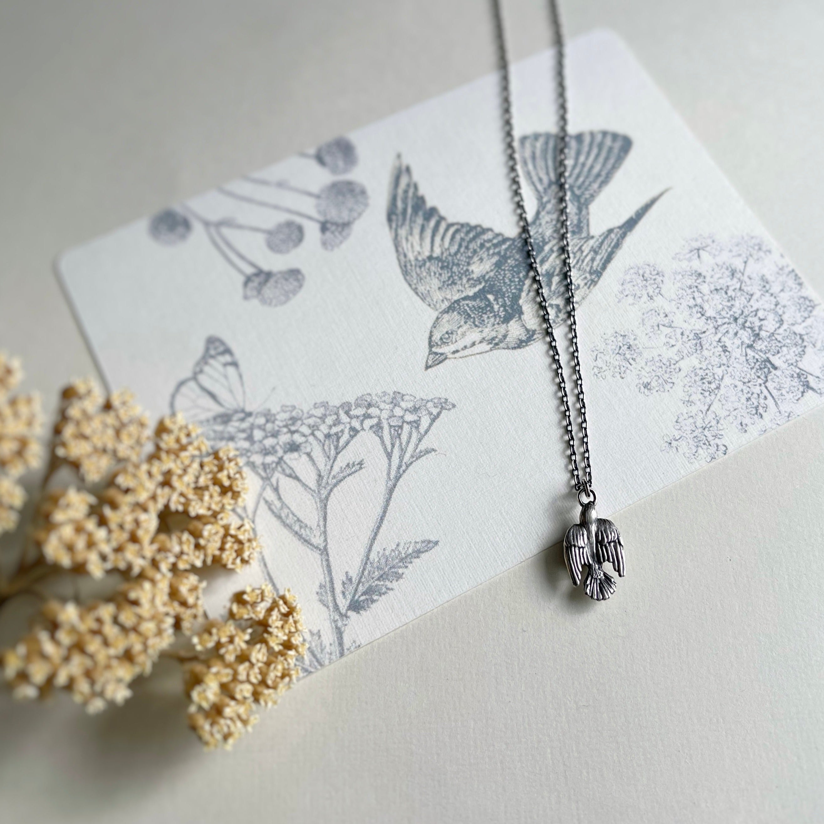 Sterling silver sparrow necklace on post card with a sparrow, wild flowers, and a butterfly, with dried yarrow